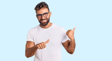 Gündelik kıyafetler giyen ve gözlüklü genç bir adam arka tarafı işaret ediyor. Elleri havada, kendinden emin bir şekilde gülümsüyor. 