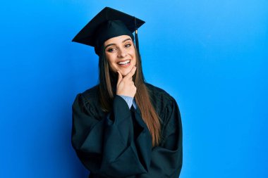 Mezuniyet şapkası ve tören cübbesi giyen güzel esmer kadın kameraya kendinden emin bakıyor kollarını kavuşturmuş ve çenesini kaldırmış gülümsüyor. Olumlu düşünmek. 