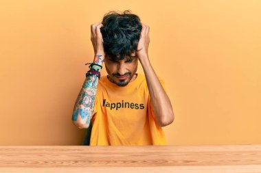 Üzerinde mutluluk mesajı olan tişört giyen genç İspanyol adam acı ve migren yüzünden çaresiz ve stresli bir şekilde masada oturuyor. Eller başının üstünde.. 