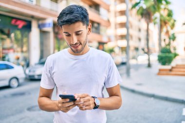 Genç Latin adam mutlu bir şekilde gülümsüyor ve şehirde akıllı telefon kullanıyor..