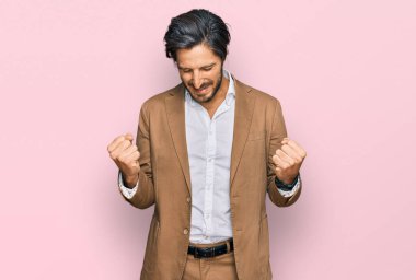 İş kıyafetleri giymiş genç İspanyol bir adam çok mutlu ve heyecanlı bir şekilde kollarını kaldırıp, gülümseyerek ve başarı için bağırarak kazanan bir jest yapıyor. kutlama konsepti. 