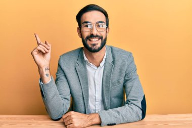 Ofiste çalışan genç İspanyol adam mutlu bir şekilde elini ve parmağını yana uzatarak gülümsüyor. 