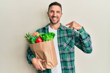 Sakallı yakışıklı bir adam, elinde poşetle market alışverişi yapıyor. Kendine güvenen, gülümseyen, kendini gururlu ve mutlu gösteren bir adam.. 