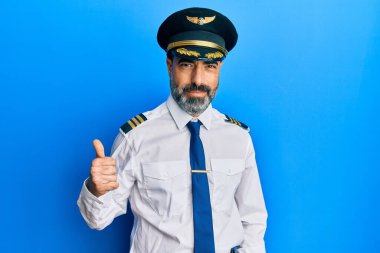 Orta yaşlı, sakallı, gri saçlı, uçak pilotu üniforması giyen mutlu bir el hareketi yapan bir adam. Kameraya bakıp başarıyı gösteren ifade. 