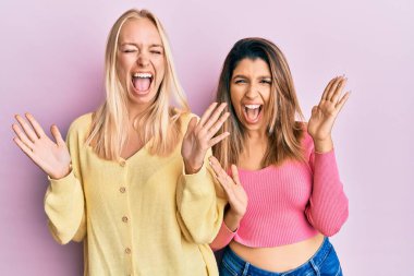 Two friends standing together over pink background celebrating mad and crazy for success with arms raised and closed eyes screaming excited. winner concept 