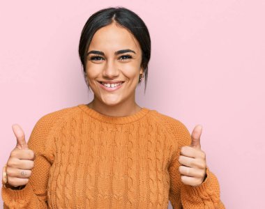 Kışlık süveter giyen genç esmer kadın elinde olumlu bir jest yapıyor, gülümsüyor ve mutlu. Neşeli ifade ve kazanan jest. 