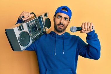 Latin asıllı genç bir adam elinde teyp, şok olmuş bir şekilde mikrofonla müzik dinliyor, şüpheci ve alaycı görünüyor, ağzı açık bir şekilde şaşırmış. 