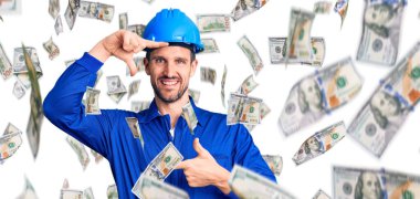 Genç, yakışıklı, işçi üniforması giyen, kasketli gülümseyen, elleri ve parmakları gülen yüzlü bir adam. yaratıcılık ve fotoğrafçılık kavramı.