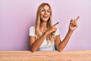 Güzel sarışın genç kadın, gündelik giysiler içinde masada oturuyor gülümsüyor ve kameraya bakıyor. İki eliyle ve parmaklarıyla kenarı işaret ediyor.. 