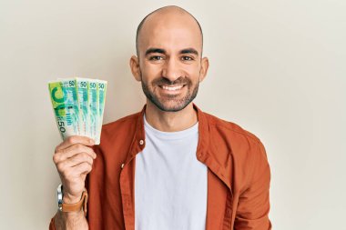 Young hispanic man holding 50 israel shekels banknotes looking positive and happy standing and smiling with a confident smile showing teeth  clipart