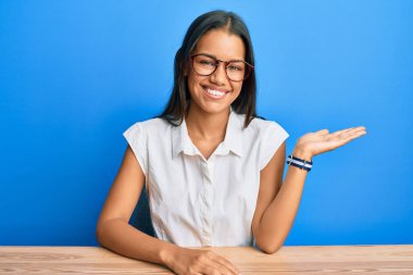 Güzel İspanyol kadın, gündelik giysiler içinde masaya oturmuş neşeli bir şekilde gülümsüyor ve avuç içiyle kameraya bakıyor.. 