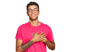 Genç, yakışıklı, Afro-Amerikalı sıradan kıyafetler giyen, elleri göğsünde, gözleri kapalı gülümseyen ve yüzünde minnet ifadesi olan bir adam. sağlık kavramı. 