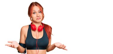 Young redhead woman wearing gym clothes and using headphones clueless and confused expression with arms and hands raised. doubt concept.  clipart