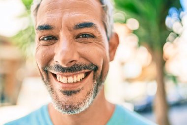 Orta yaşlı, kır saçlı, gülümseyen bir adam şehrin caddesinde mutlu bir şekilde yürüyor..