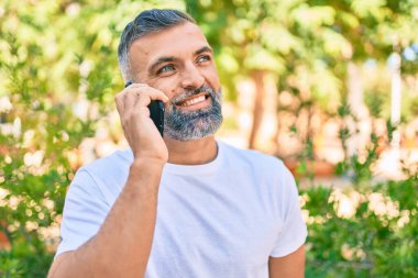 Orta yaşlı, kır saçlı, gülümseyen, mutlu, parkta akıllı telefondan konuşan bir adam.