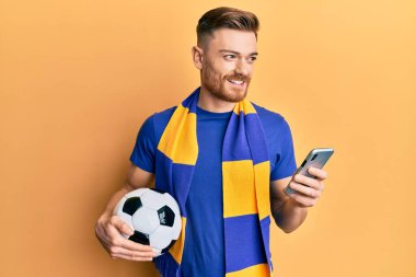 Genç kızıl saçlı adam holigan atkısı takıyor ve akıllı telefon kullanıyor. Gülümsüyor, yan tarafa bakıyor ve düşünüyor.. 