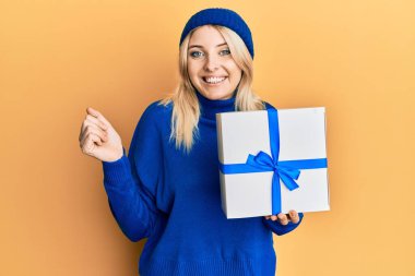 Young caucasian woman holding gift screaming proud, celebrating victory and success very excited with raised arm 
