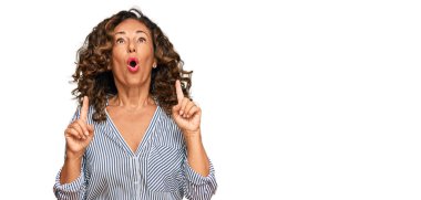 Orta yaşlı İspanyol kadın gündelik kıyafetler giyip şaşkın şaşkın yukarı bakıyor ve parmaklarıyla işaret ediyor ve kollarını kaldırıyor.. 