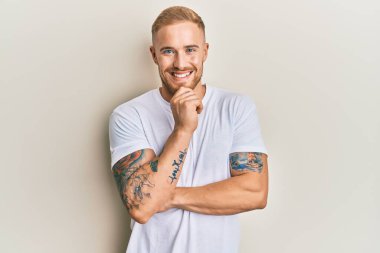 Gündelik beyaz tişört giyen genç beyaz bir adam kameraya kollarını kavuşturmuş ve çenesinde elle bakıyor. Olumlu düşünmek. 