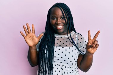 Saçları örgülü genç siyahi bir kadın 7 numarayı gösterip gösterirken kendinden emin ve mutlu bir şekilde gülümsüyor.. 