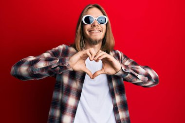Handsome caucasian man with long hair wearing hipster shirt wearing sunglasses smiling in love doing heart symbol shape with hands. romantic concept. 