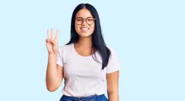 Genç, güzel Asyalı bir kız günlük kıyafetler ve gözlük takıyor. Üç numaralı parmaklarıyla gösteriyor ve işaret ediyor. Kendinden emin ve mutlu bir şekilde gülümsüyor.. 