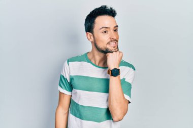 Elleri çenesinde gündelik giysiler içinde soru soran genç İspanyol bir adam, düşünceli bir ifade. Gülümseyen ve düşünceli bir yüz. şüphe kavramı. 