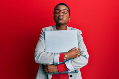 Genç Afro-Amerikalı iş kadını dizüstü bilgisayarını elinde tutuyor ve komik suratıyla yanaklarını şişiriyor. Ağzı havayla şişer, havayı yakalar.. 