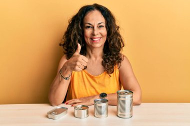 Beautiful middle age woman with canned food smiling happy and positive, thumb up doing excellent and approval sign 