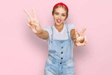 Kafkasyalı genç bir kız günlük kıyafetler giymiş, dili dışarı fırlamış bir şekilde gülümsüyor ve iki elini de zafer işareti yapıyor. İki numara.. 