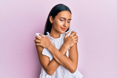 Zarif bir yaz gömleği giyen genç esmer kadın kendine sarılıyor mutlu ve pozitif, kendinden emin gülümsüyor. Sevgi ve özgüven. 