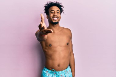Young african american man with beard wearing swimsuit smiling friendly offering handshake as greeting and welcoming. successful business. 