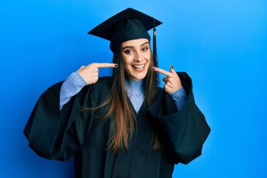 Mezuniyet şapkası ve tören cübbesi takan güzel esmer bir kadın neşeli bir şekilde gülümseyip, parmaklarıyla ve ağzıyla işaret ediyor. diş sağlığı kavramı. 