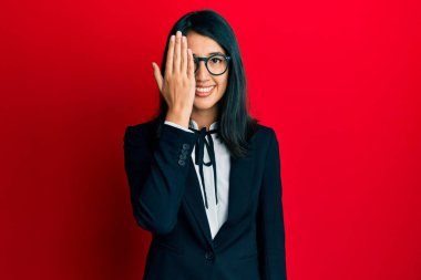 Güzel Asyalı genç bayan iş elbisesi giyiyor. Bir gözünü eliyle kapatıyor. Yüzünde kendine güvenen bir gülümseme ve sürpriz bir duygu var.. 