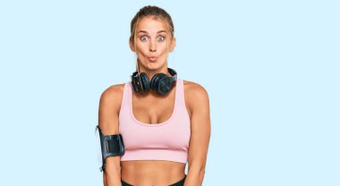 Young blonde woman wearing gym clothes and using headphones making fish face with lips, crazy and comical gesture. funny expression. 