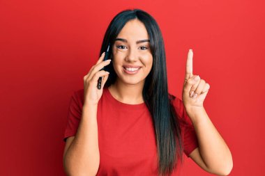 Young beautiful hispanic girl having conversation talking on the smartphone smiling with an idea or question pointing finger with happy face, number one 