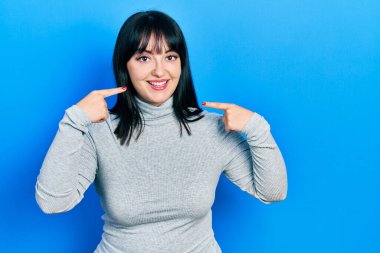 Günlük kıyafetler giyen, neşeli gülümseyen, parmaklarıyla ve ağzıyla işaret eden genç İspanyol kadın. diş sağlığı kavramı. 