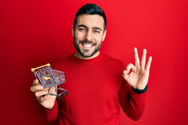 Young hispanic man holding small supermarket shopping cart doing ok sign with fingers, smiling friendly gesturing excellent symbol 