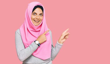 Geleneksel İslami başörtüsü takan genç beyaz kadın gülümseyip kameraya bakıyor ve iki eliyle ve parmaklarıyla kenarı işaret ediyor.. 