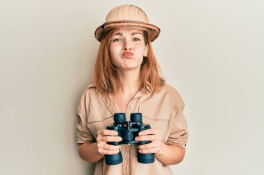 Genç beyaz kadın kaşif şapkası takıyor dürbünle garip bir yüz ifadesi takınarak yanaklarını şişiriyor. Ağzı havayla şişer, havayı yakalar.. 