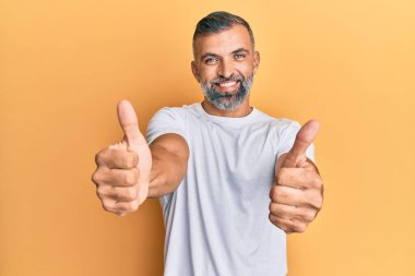 Orta yaşlı beyaz tişört giyen yakışıklı bir adam elinle olumlu jest yapmayı onaylıyor, gülümsüyor ve başarı için mutlu. kazanan jest. 