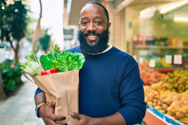Sakallı Afrikalı Amerikalı bir adam elinde süpermarketten alınmış kağıt poşetler tutuyor.