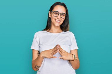 Latin kökenli genç bir kadın günlük beyaz tişörtler giyiyor ellerini birleştirip gülümseyerek gevşemiş ve neşeli olmasını diliyordu. Başarı ve iyimserlik 