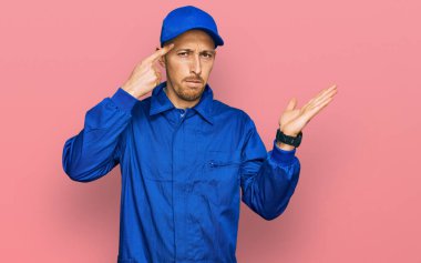 Bald man with beard wearing builder jumpsuit uniform confused and annoyed with open palm showing copy space and pointing finger to forehead. think about it. 