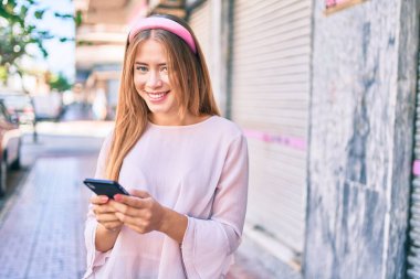 Genç beyaz kız şehirde akıllı telefon kullanarak mutlu bir şekilde gülümsüyor..