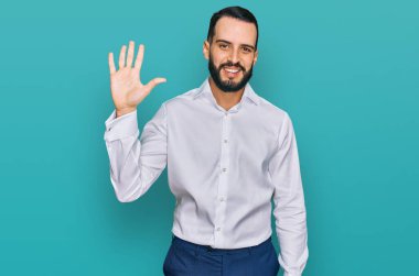 Sakallı genç adam iş gömleği giyiyor ve parmaklarıyla beş numarayı gösterirken kendinden emin ve mutlu bir şekilde gülümsüyor.. 