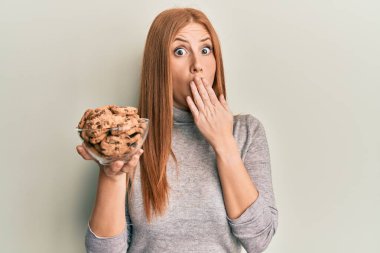 Genç İrlandalı kadın elinde çikolata parçacıklı kurabiyelerle ağzı elle kaplanmış, şok olmuş ve hata yapmaktan korkan. şaşırmış ifade 