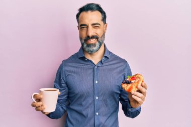 Orta yaşlı, sakallı, kır saçlı kahve içen ve pasta yiyen bir adam kameraya seksi bir ifadeyle bakıyor, neşeli ve mutlu bir yüzle.. 