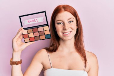 Genç kızıl saçlı kız, elinde allık renginde bir palet tutuyor. Pozitif ve mutlu görünüyor. Kendine güvenen bir gülümsemeyle dişlerini gösteriyor. 