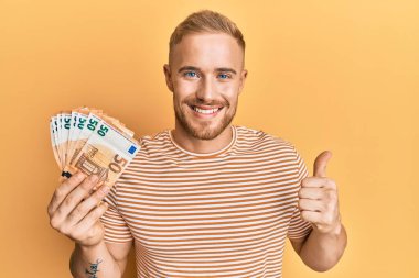 Elinde 50 Euro 'luk banknotlar tutan genç beyaz adam mutlu ve olumlu gülümsüyor, başparmağıyla mükemmel ve onay işareti yapıyor. 